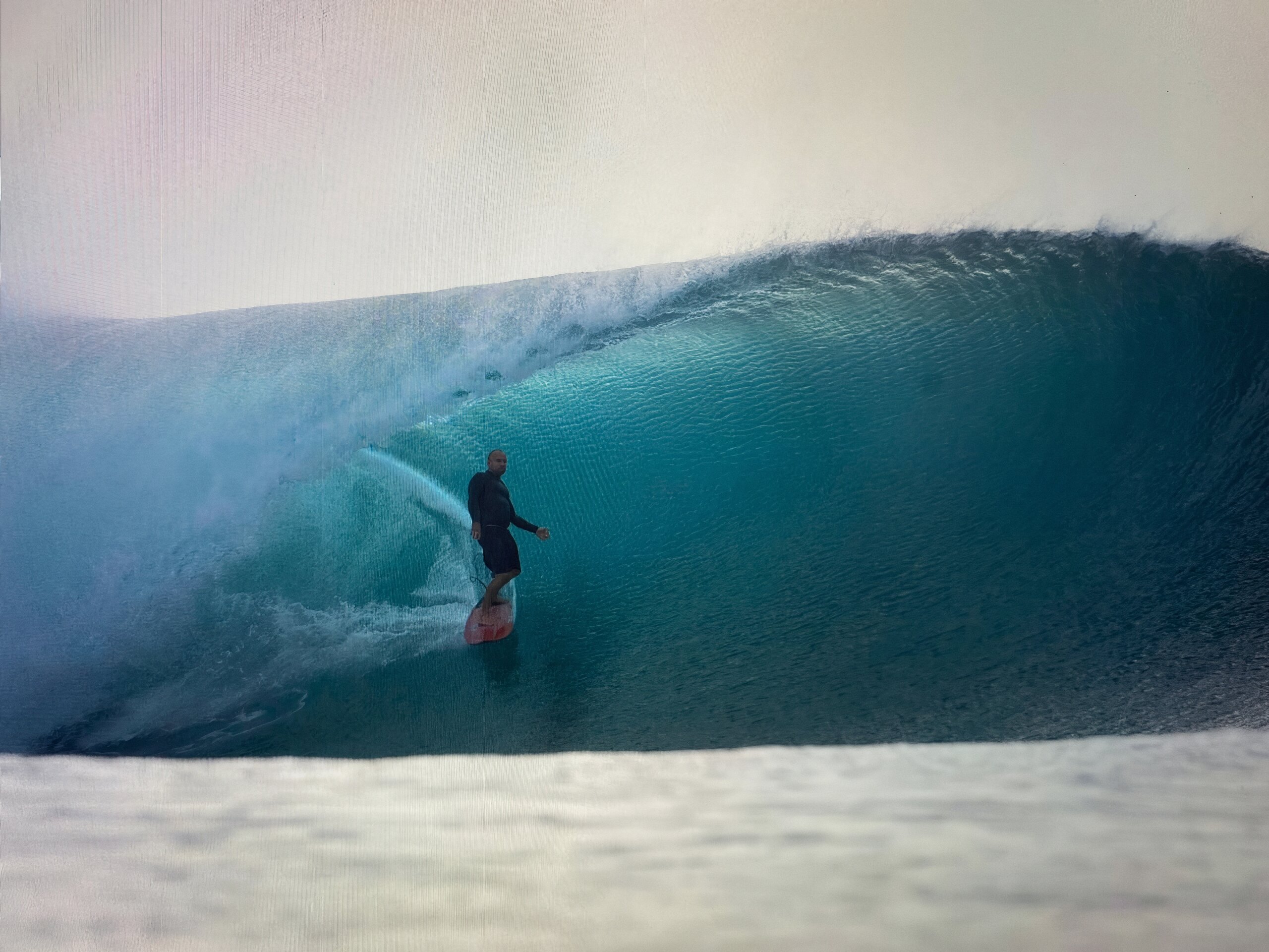 A surfer in a yellow helmet drops in on a large, clean ocean wave — the wave standing tall and turquoise, the rider committed at the take-off, a long open face below
