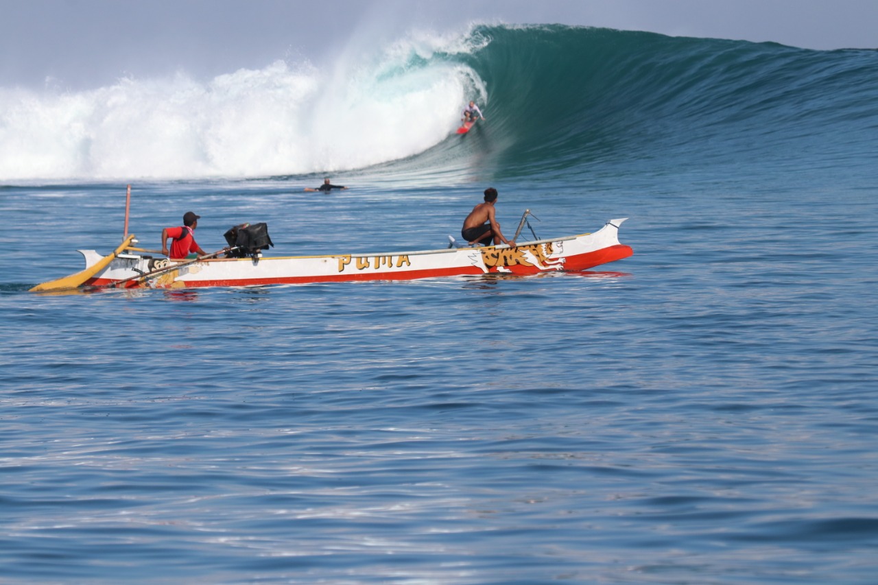 A traditional Indonesian outrigger painted red and white with the name PUMA on the hull, two local boatmen aboard, a surfer riding inside the barrel of a clean wave in the background