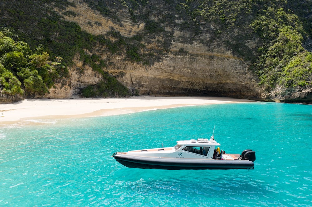 Kai Koa at anchor in a turquoise bay below the limestone cliffs of the Nusa islands — a thirteen-metre centre-console surf charter boat with hardtop and outboards, the water clear enough to read the sand under the keel
