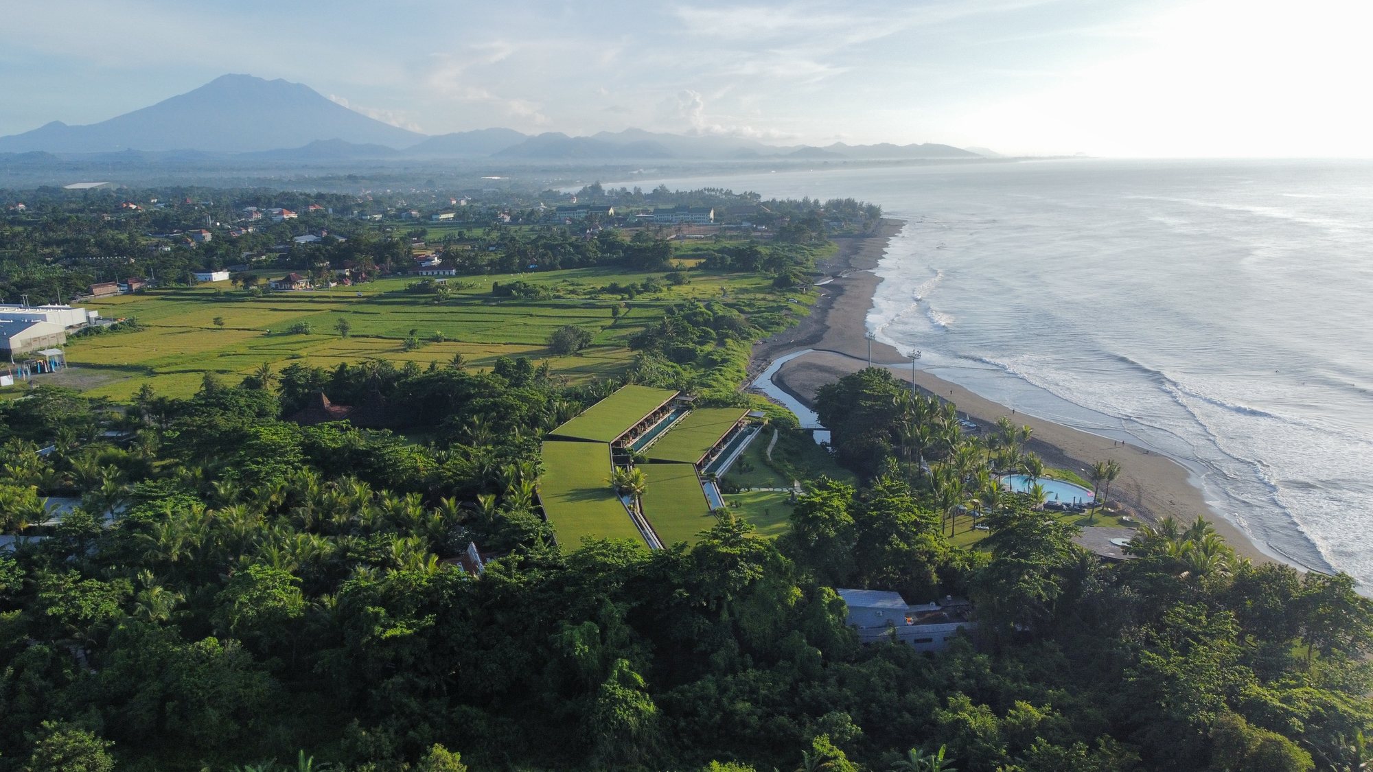 Aerial view of Komune Bali at first light — the property with green rooftop lawns sited above Keramas beach, Mount Agung rising in the distance, rice fields and the river mouth in the middle distance