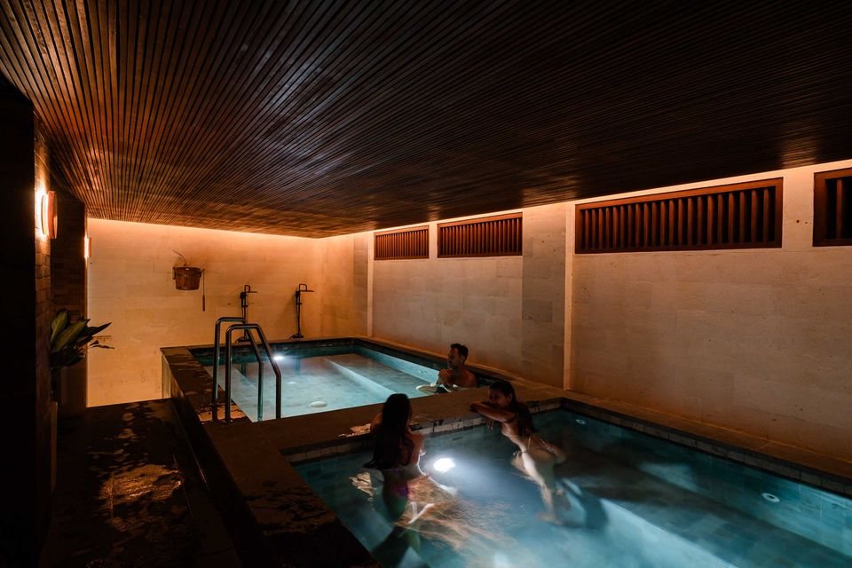 The magnesium spa at the Komune recovery centre — two stone pools in an indoor sandstone-walled room, slatted teak ceiling, low warm lighting