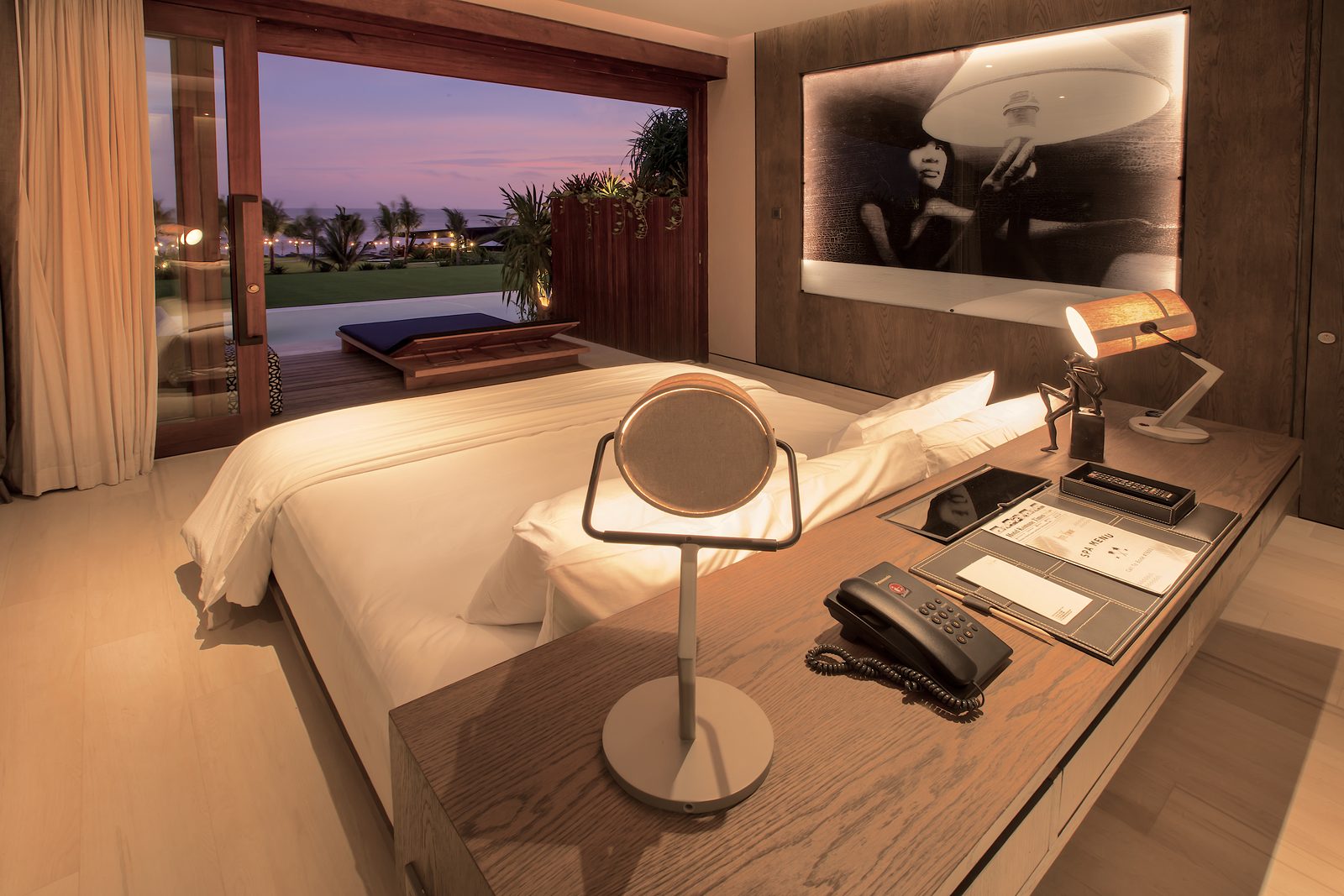 A Komune suite at dusk — king bed on a low oak platform, large backlit photographic artwork in the headboard wall, floor-to-ceiling sliding doors open to a private pool terrace and the ocean beyond at sunset