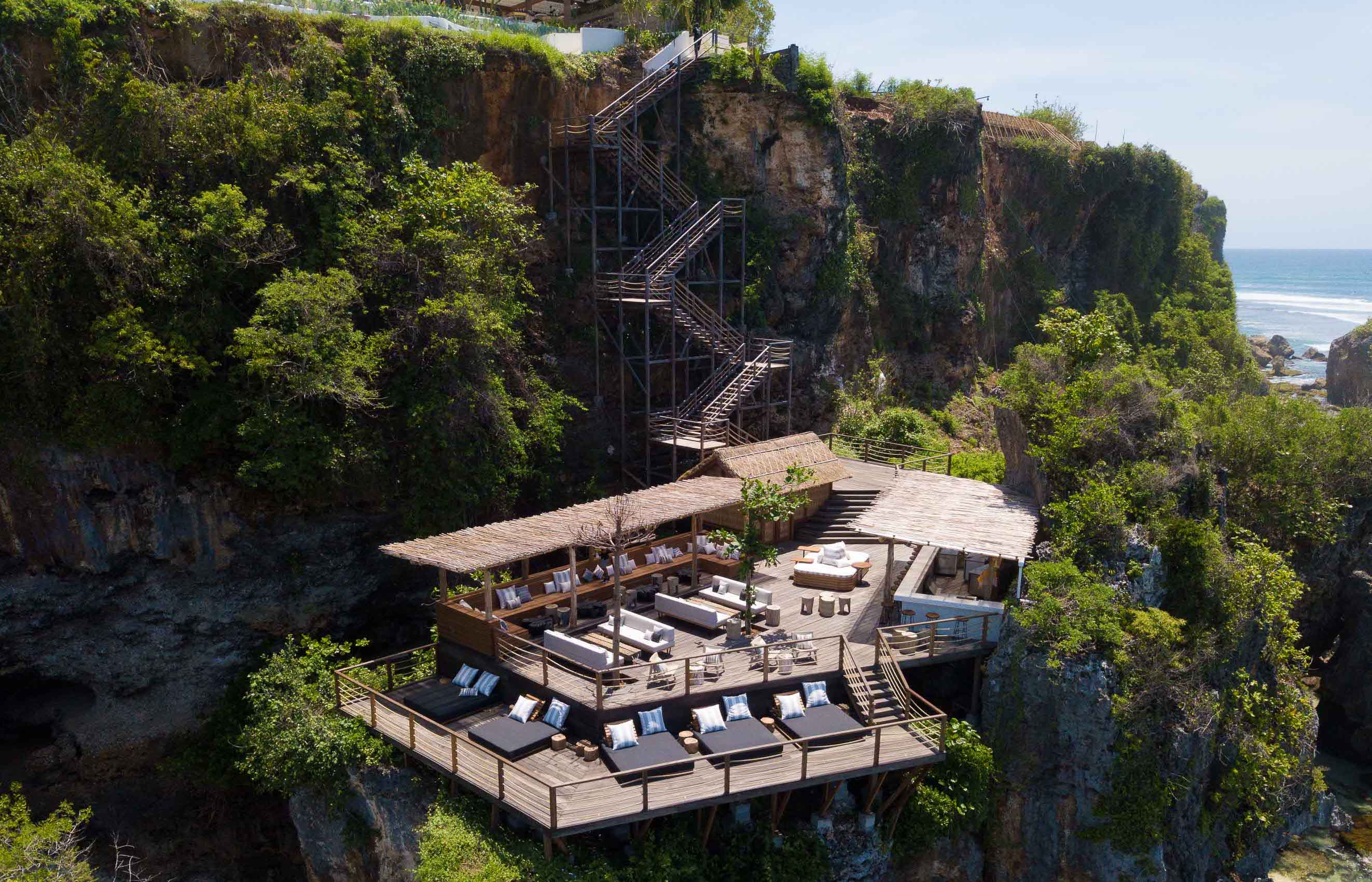 The Ocean Deck at Ulu Cliffhouse — bleached timber decking and a bar set into the rock face below the main venue, the staircase climbing the cliff to the clifftop, the Indian Ocean break to the right