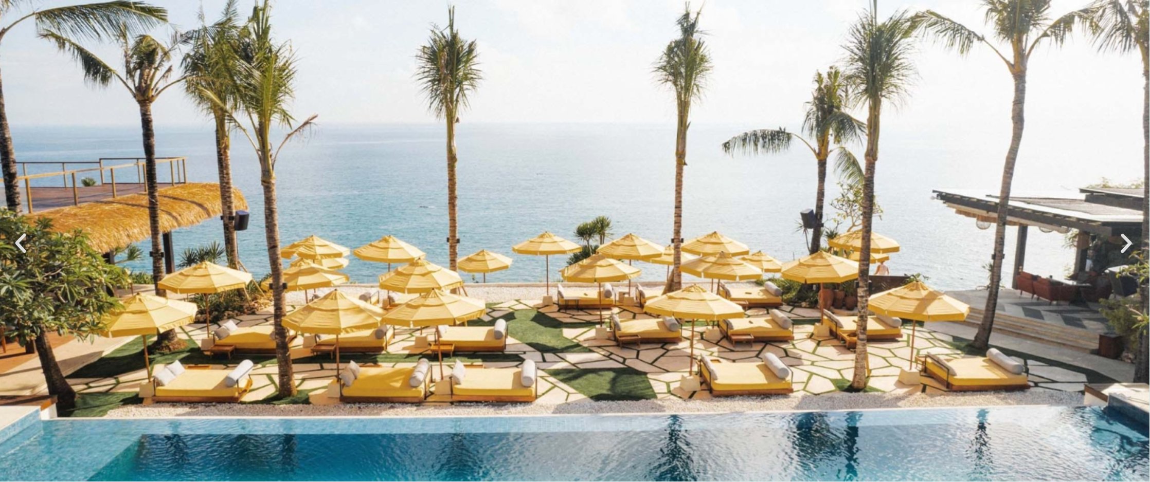 The clifftop pool deck at Ulu Cliffhouse — rows of yellow umbrellas and daybeds laid out across a terrace of grass-and-flagstone, palm trees rising between them, the Indian Ocean stretching to the horizon, the rock-bar pavilion to the right and the upper-cliff platform to the left