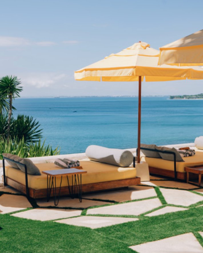 Two yellow daybeds under a single yellow umbrella on the Ulu Cliffhouse pool terrace — palm fronds to the left, the green-and-flagstone deck underfoot, the Indian Ocean and the southern coastline of the Bukit in the distance