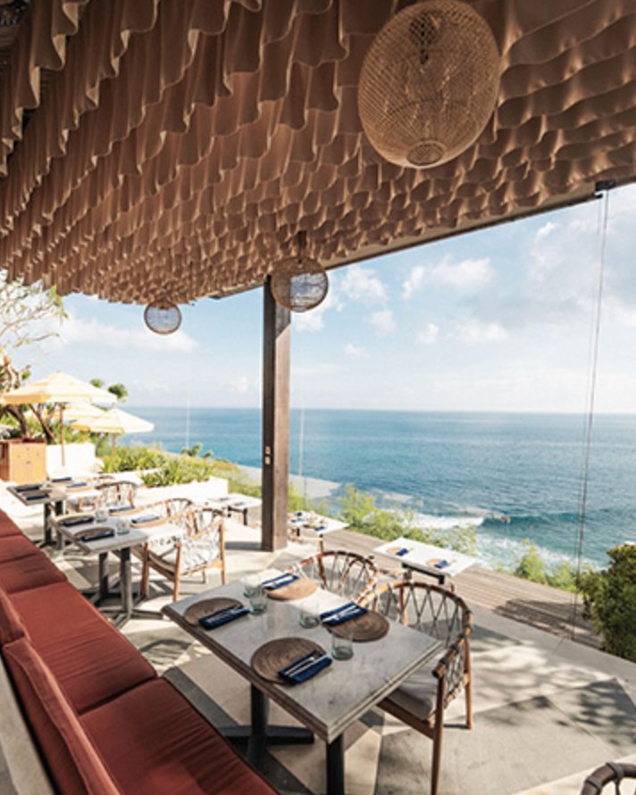 The upper-deck restaurant at Ulu Cliffhouse — a sculptural draped fabric ceiling in warm sand tones above marble tables set for service, wicker dining chairs, a rust-coloured banquette in the foreground, the open frame looking out across the deck to the Indian Ocean