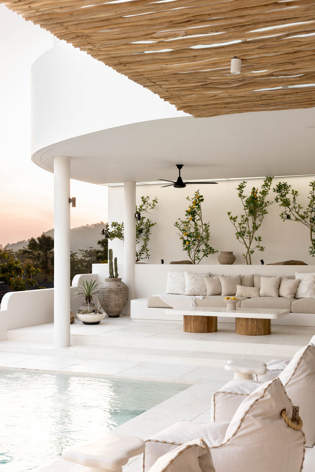 The pool terrace at Villa Zyloh — citrus trees in old terracotta urns, white linen sofas under a woven driftwood pergola, the salt-water pool in the foreground